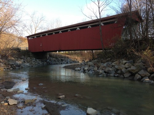 A red covered bridge spans a calm stream, surrounded by bare trees and rocky banks under a clear sky.