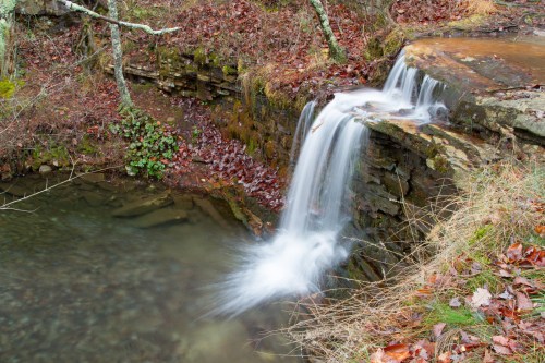 A serene waterfall cascades over rocky ledges into a calm pool, surrounded by autumn leaves and greenery.