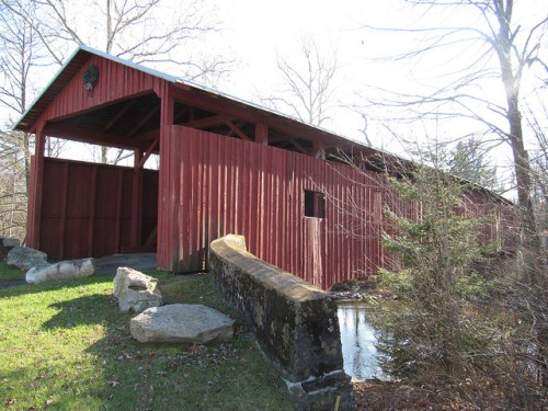 A red covered bridge stands beside a stone wall, surrounded by trees and grass on a sunny day.