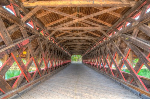 A wooden covered bridge interior with a crisscross pattern and a view of greenery at the far end.