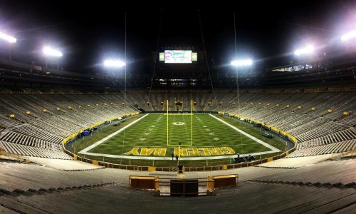 A wide view of an empty football stadium at night, with bright lights illuminating the field and seating.