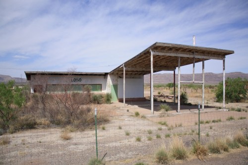 Abandoned building with a large overhang, surrounded by sparse vegetation and mountains in the background.