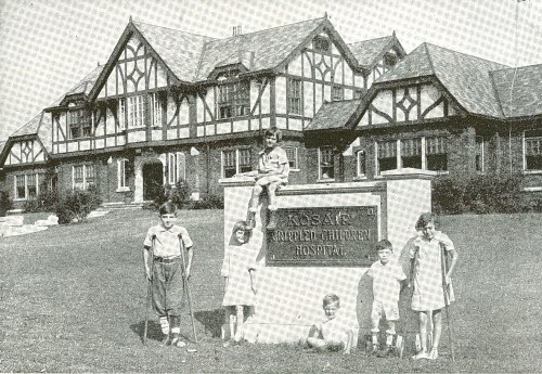 A group of children poses in front of a historic building, labeled "Tobat Hospital," on a grassy lawn.