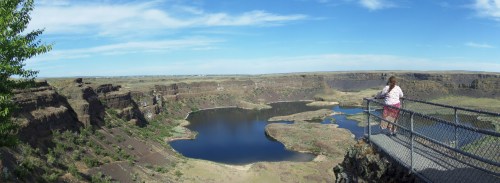 A person stands on a viewing platform overlooking a vast canyon and lake under a blue sky with scattered clouds.