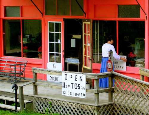 A woman in a blue skirt adjusts a sign outside a pink storefront with open and closed hours displayed.