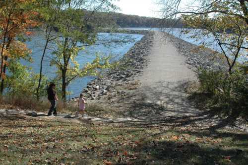 A woman and a child walk along a path by a river, with trees and autumn leaves in the background.