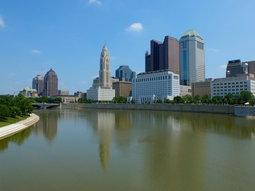 A skyline view of a city with tall buildings reflecting in a river under a clear blue sky.