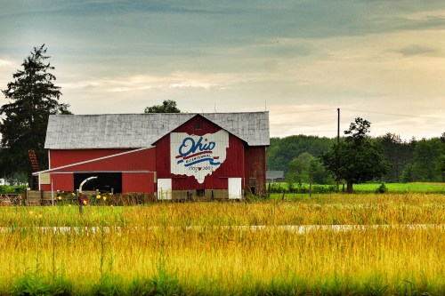 Red barn with "Ohio" logo, surrounded by tall grass and trees under a cloudy sky.