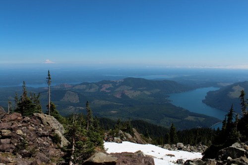 A panoramic view of mountains and a lake under a clear blue sky, with snow patches and distant peaks visible.