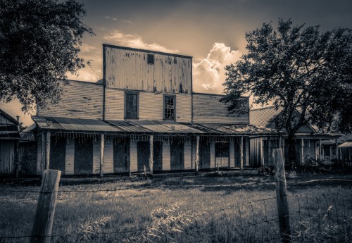 Black and white image of an old, weathered building with a rusty roof and boarded windows, surrounded by grass and trees.