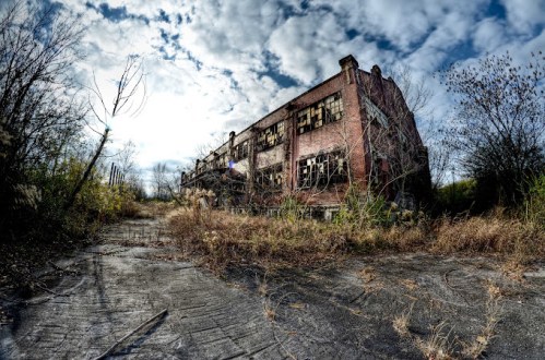 Abandoned brick building surrounded by overgrown vegetation and a cloudy sky.