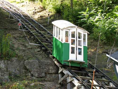 A green and white funicular car ascends a steep, wooded hill on a set of tracks.