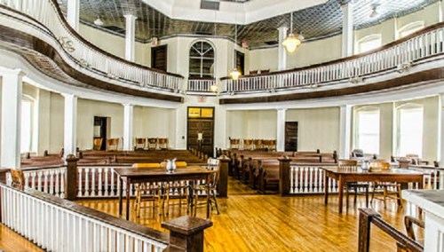Interior of a historic courtroom with wooden benches, tables, and a balcony, featuring large windows and a polished floor.