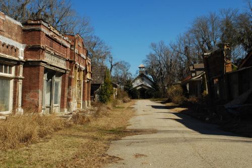 Abandoned buildings line a deserted road, surrounded by overgrown grass and bare trees under a clear blue sky.