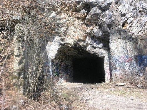 A dark cave entrance surrounded by rocky terrain and sparse vegetation, with graffiti on the stone walls.