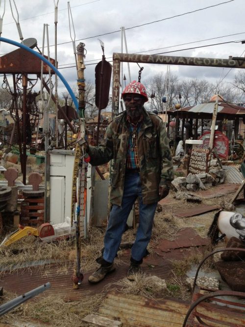 A man in a camouflage jacket and red hat stands among various metal sculptures and art in an outdoor setting.