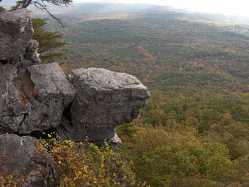 A rocky outcrop overlooks a vast, colorful forested landscape under a cloudy sky.