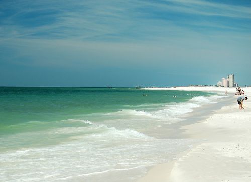 A serene beach scene with gentle waves, soft white sand, and a distant skyline under a blue sky.