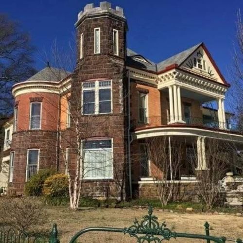 Victorian-style house with a turret, red brick exterior, and a wraparound porch surrounded by trees and shrubs.