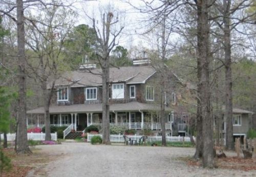 A large, two-story house surrounded by trees, with a gravel driveway and a white picket fence.