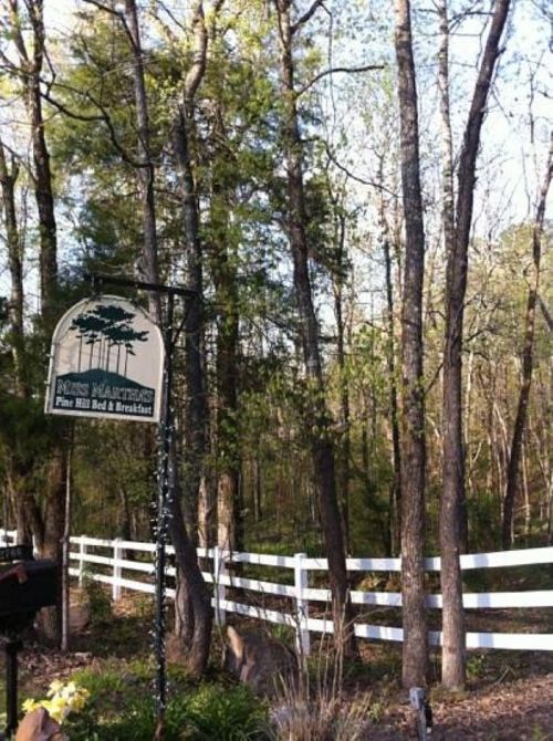 Sign for Pine Hill Bed & Breakfast surrounded by trees and a white fence in a wooded area.