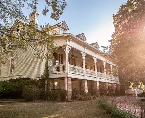 A large, historic house with a wraparound porch, surrounded by trees and bathed in warm sunlight.