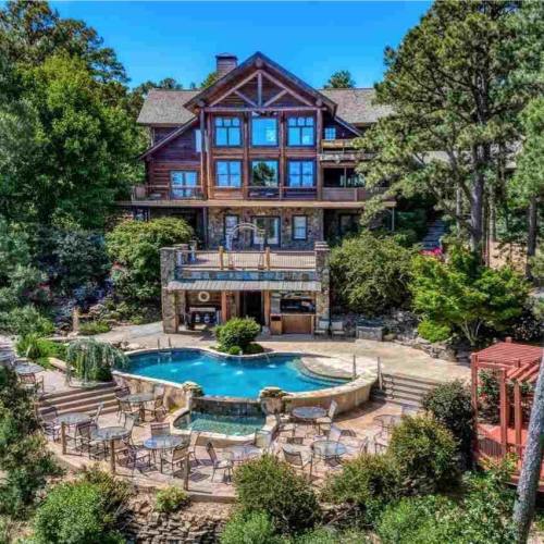 A spacious backyard with a pool, hot tub, and seating area, surrounded by lush greenery and a large wooden house.