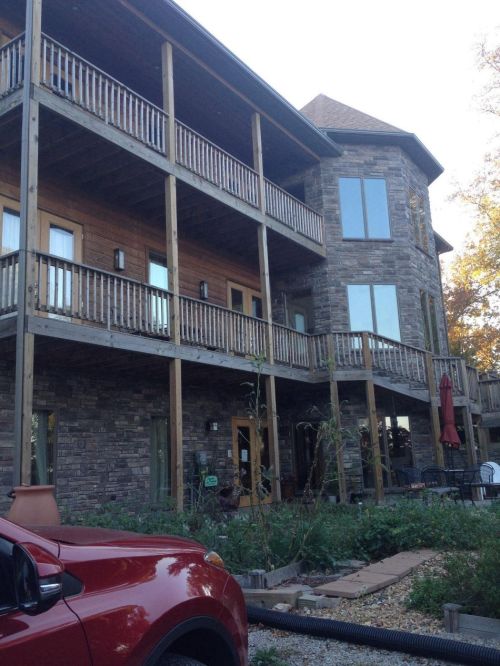 A multi-level stone house with wooden balconies, surrounded by greenery and a red car parked in front.