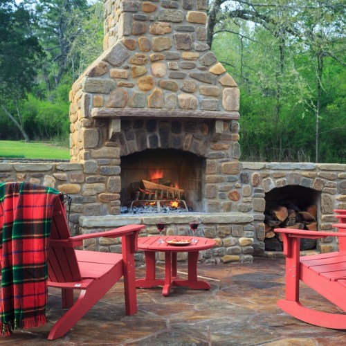 Cozy outdoor seating area with a stone fireplace, red chairs, a plaid blanket, and two glasses of wine on a table.
