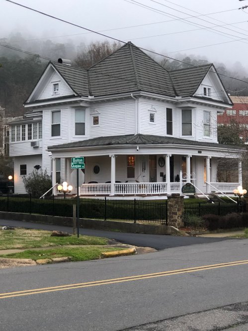 A large white Victorian-style house with a wraparound porch, surrounded by a black fence and street signs nearby.