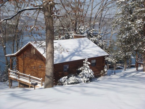 A cozy wooden cabin covered in snow, surrounded by trees and overlooking a serene lake.