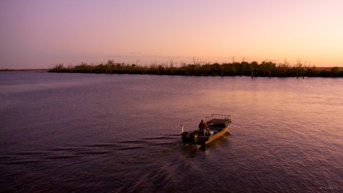 A small boat glides across a calm river at sunset, with trees silhouetted against a colorful sky.