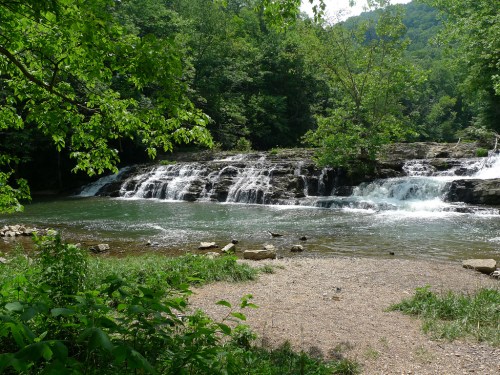 A serene waterfall cascading over rocks into a calm river, surrounded by lush green trees and mountains in the background.