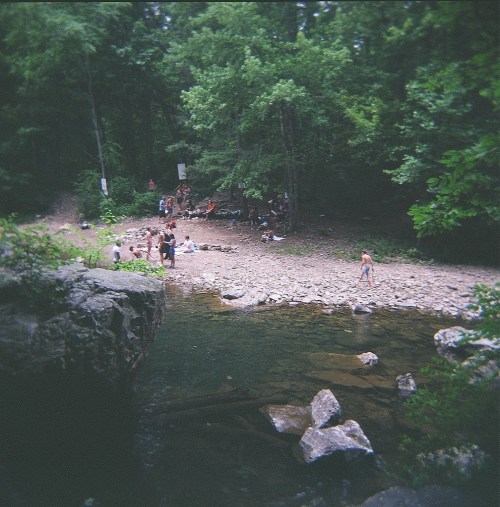 A serene forest scene with people gathered by a rocky riverbank, surrounded by lush greenery and trees.