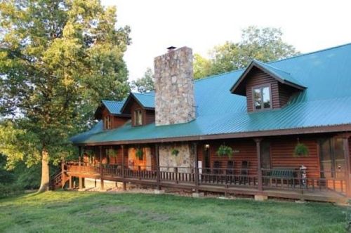 A rustic wooden cabin with a stone chimney, surrounded by trees and a grassy area, featuring a porch with chairs.