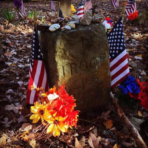 A grave marker with flags, flowers, and stones, surrounded by fallen leaves in a serene outdoor setting.