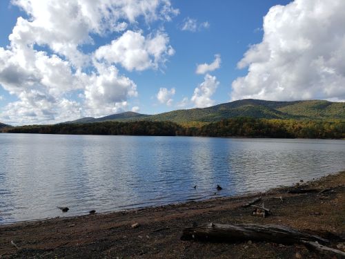 A serene lake surrounded by mountains, with fluffy clouds in a blue sky and a sandy shore.