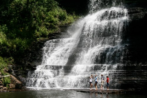 Three people stand near a cascading waterfall surrounded by lush greenery. Water splashes down the rocky surface.