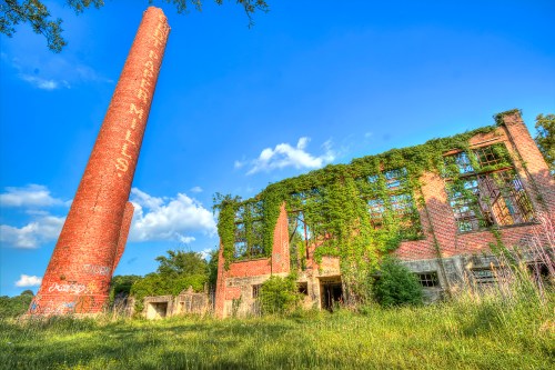 Abandoned brick mill with a tall chimney, overgrown with vines, set against a blue sky and grassy foreground.