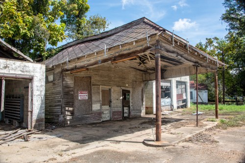 Abandoned building with a weathered roof, wooden structure, and overgrown surroundings under a clear blue sky.