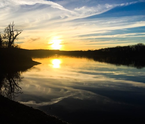 Sunset over a calm lake, with vibrant colors reflecting on the water and silhouetted trees along the shore.