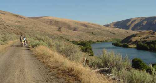 Two people ride bicycles along a dirt path beside a river, surrounded by rolling hills and sparse vegetation.