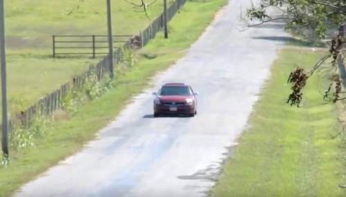 A red car drives down a rural road lined with grass and fences on a sunny day.