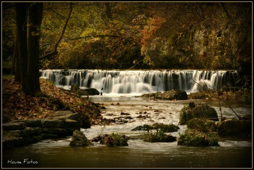 A serene waterfall cascades over rocks, surrounded by autumn foliage and a calm river.