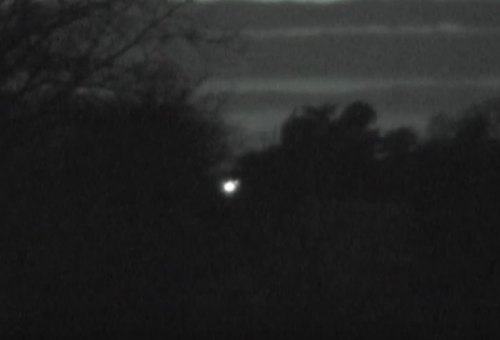 A dim light glows in the distance, surrounded by dark trees and a cloudy night sky.