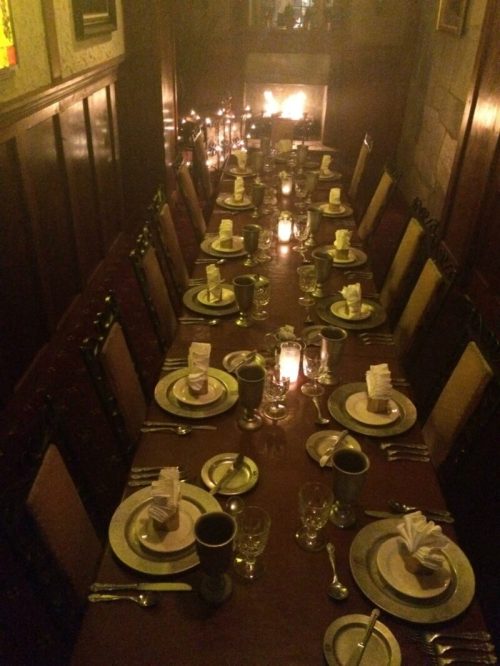 A long, elegantly set dining table with candles and a fireplace in a cozy, dimly lit room.