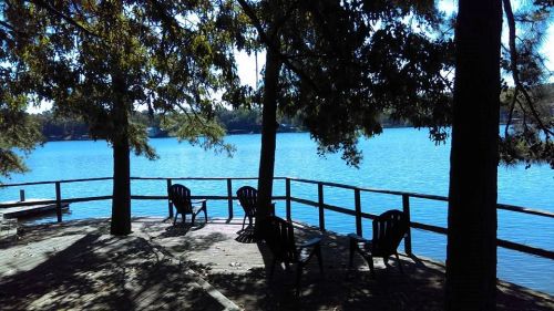 A serene lakeside view with wooden chairs on a deck, surrounded by trees and calm blue water.