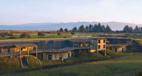 Aerial view of a modern building complex surrounded by green fields and mountains in the background during sunset.