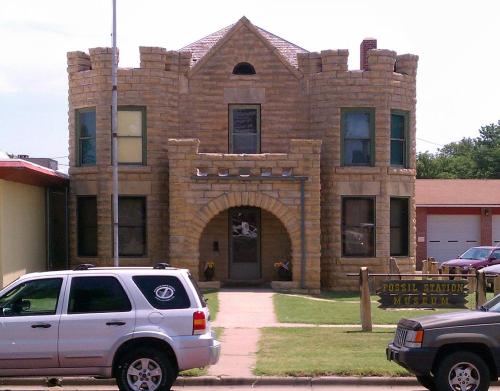 A stone castle-like building with turrets, featuring large windows and a welcoming entrance, surrounded by grass and parked cars.