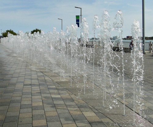 A row of water fountains spraying in a paved area near a waterfront, with a clear sky in the background.